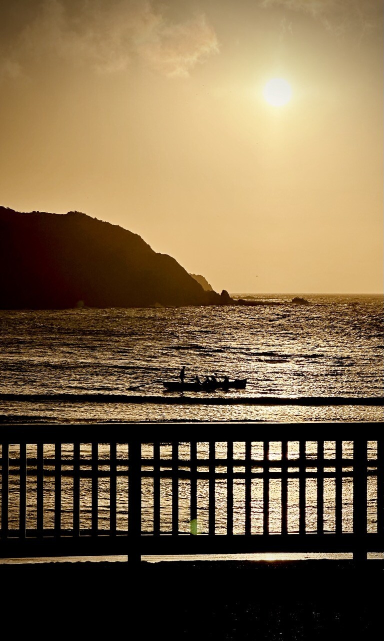 A sepia toned view across the bay, with the sun starting to come down, a boat with rowers is paused in the middle, with a fence in the foreground 