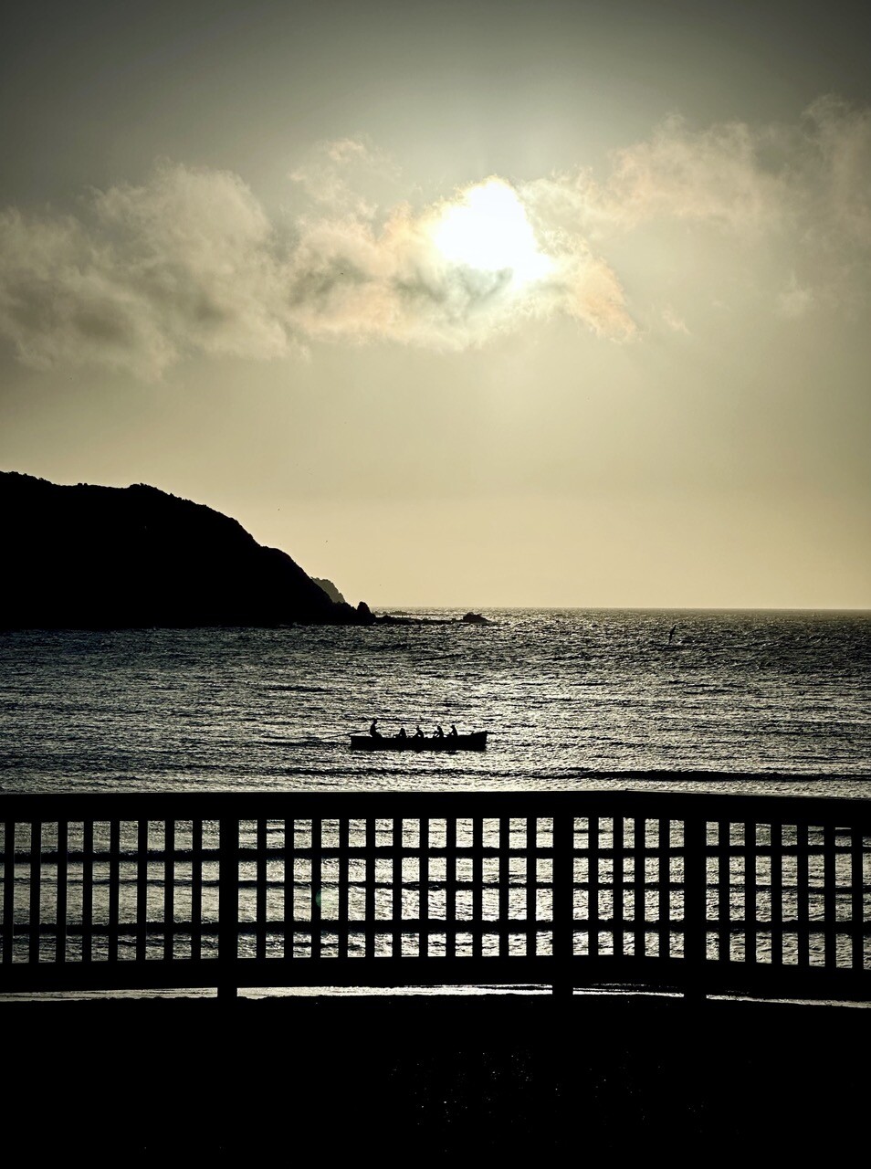 A blue/gray toned view across the bay, with the sun starting to come down, a boat with rowers is in the middle, with a fence in the foreground 